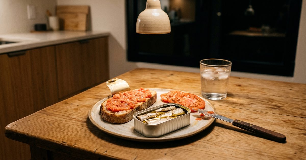 Cena montada sobre la mesa de la cocina de noche: tostada con tomate, una lata de sardinas abierta, unas rodajas de tomate y un vaso de agua