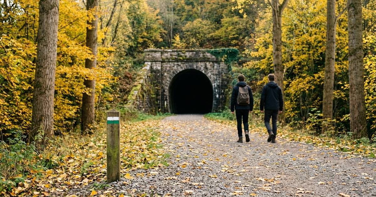 Two walkers seen from behind on a gravel greenway heading towards the stone mouth of an old railway tunnel, among autumn trees, next to a wooden post with the white-and-green local trail mark