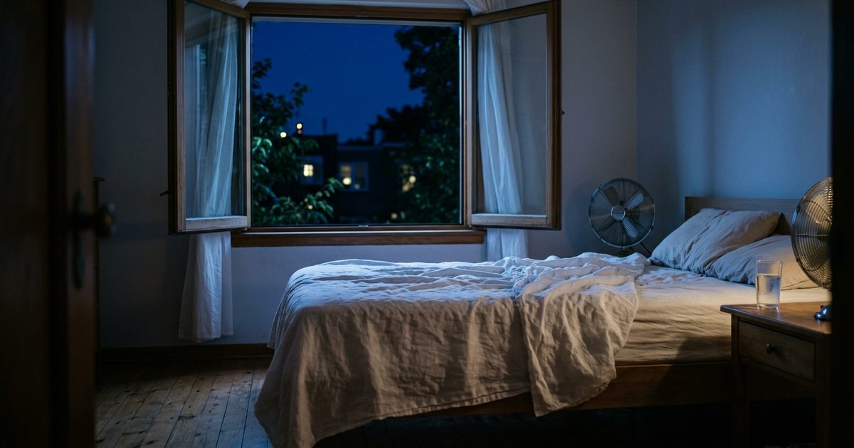 Dormitorio de noche con la ventana abierta al cielo azul oscuro, la sábana fina revuelta, un ventilador junto a la cama y un vaso de agua en la mesilla